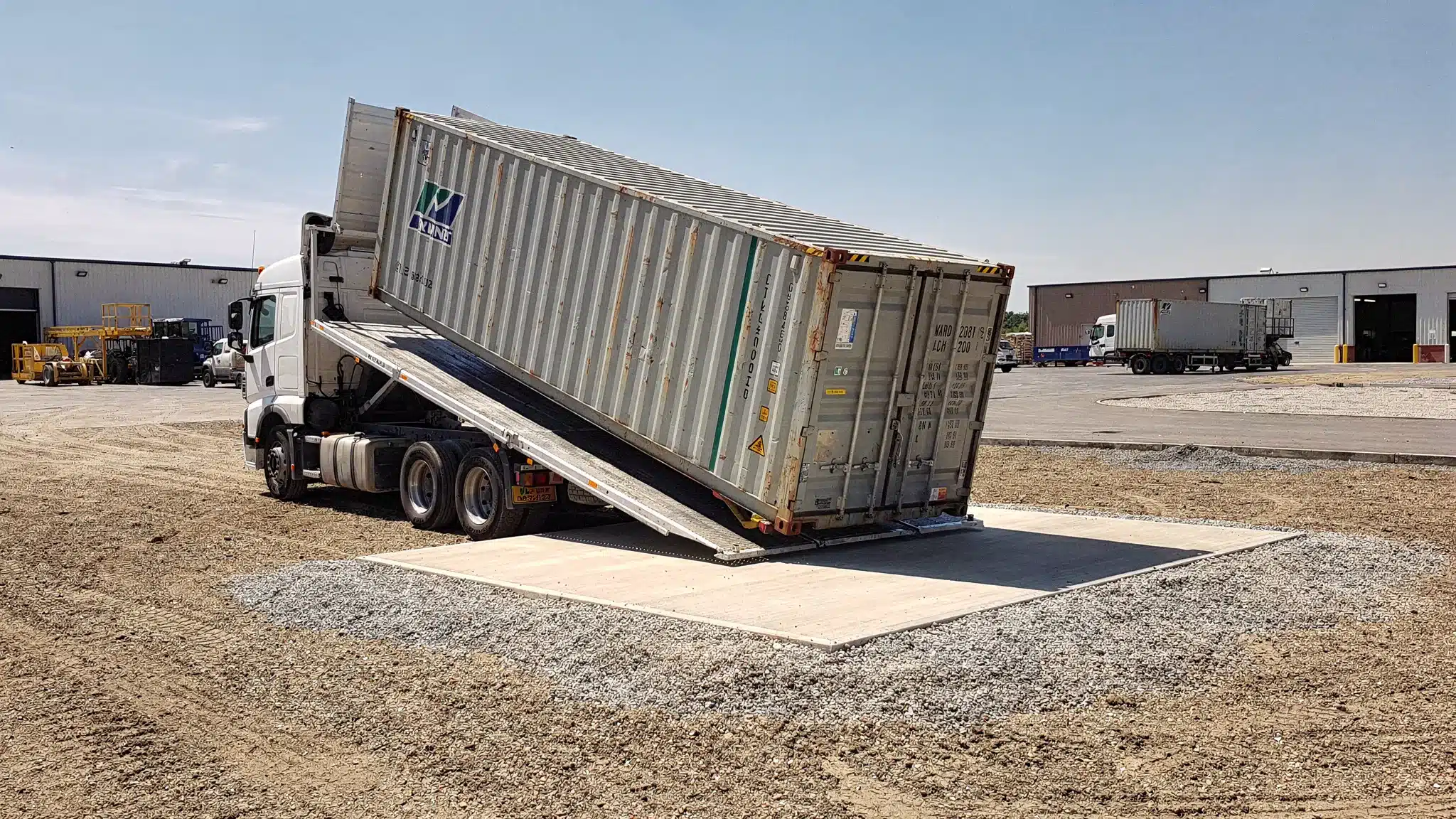 Container One Shipping Containers: Specs, Grades, Delivery 1 A shipping container being delivered on a tilt-bed truck onto a level gravel pad at a commercial site, with clear access space and no overhead power lines.