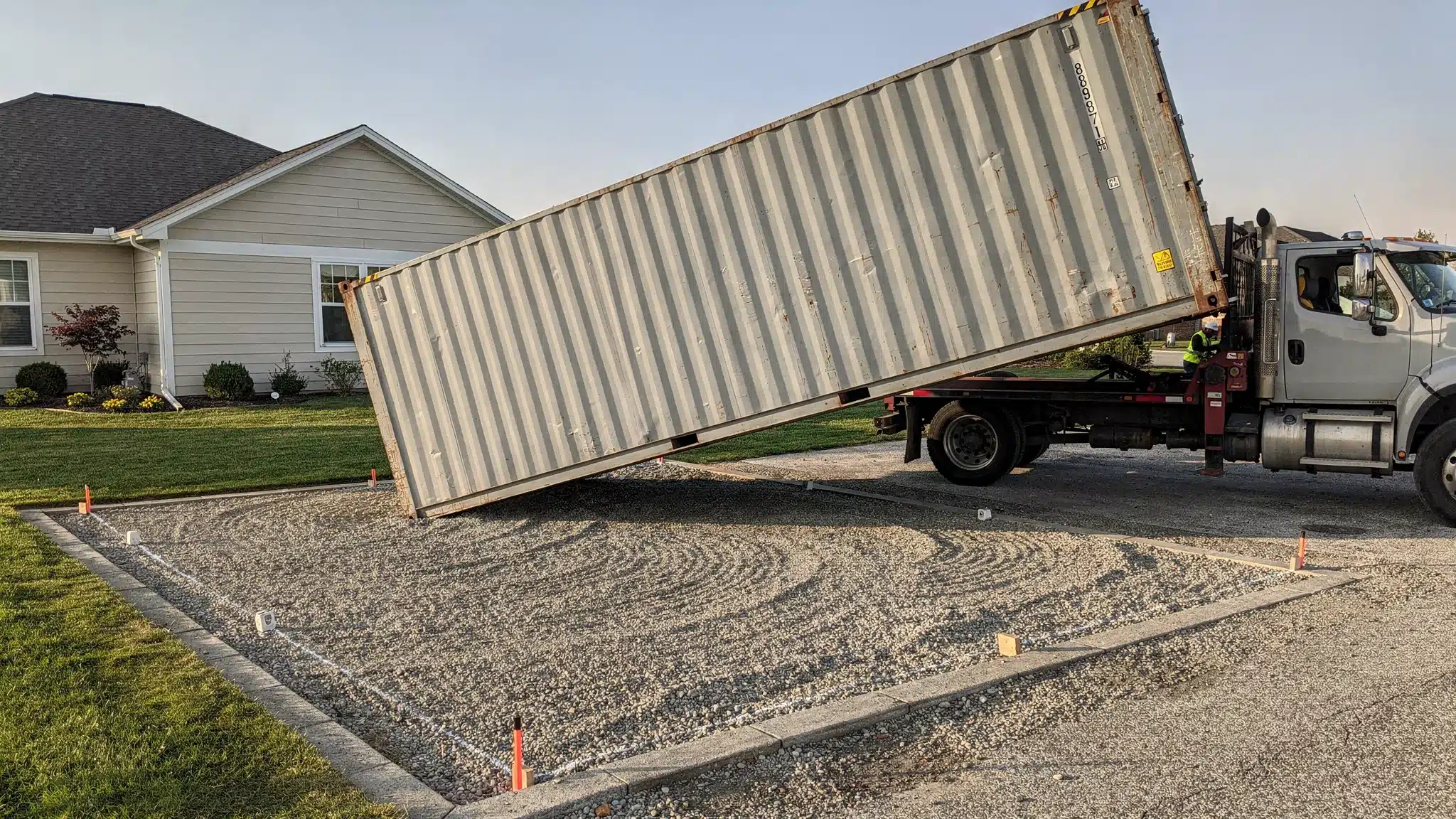 Transporting a Shipping Container: Permits, Costs, Timelines 1 A tilt-bed delivery truck placing a 20ft shipping container onto a prepared gravel pad at a residential property, with clear overhead space and marked placement corners.