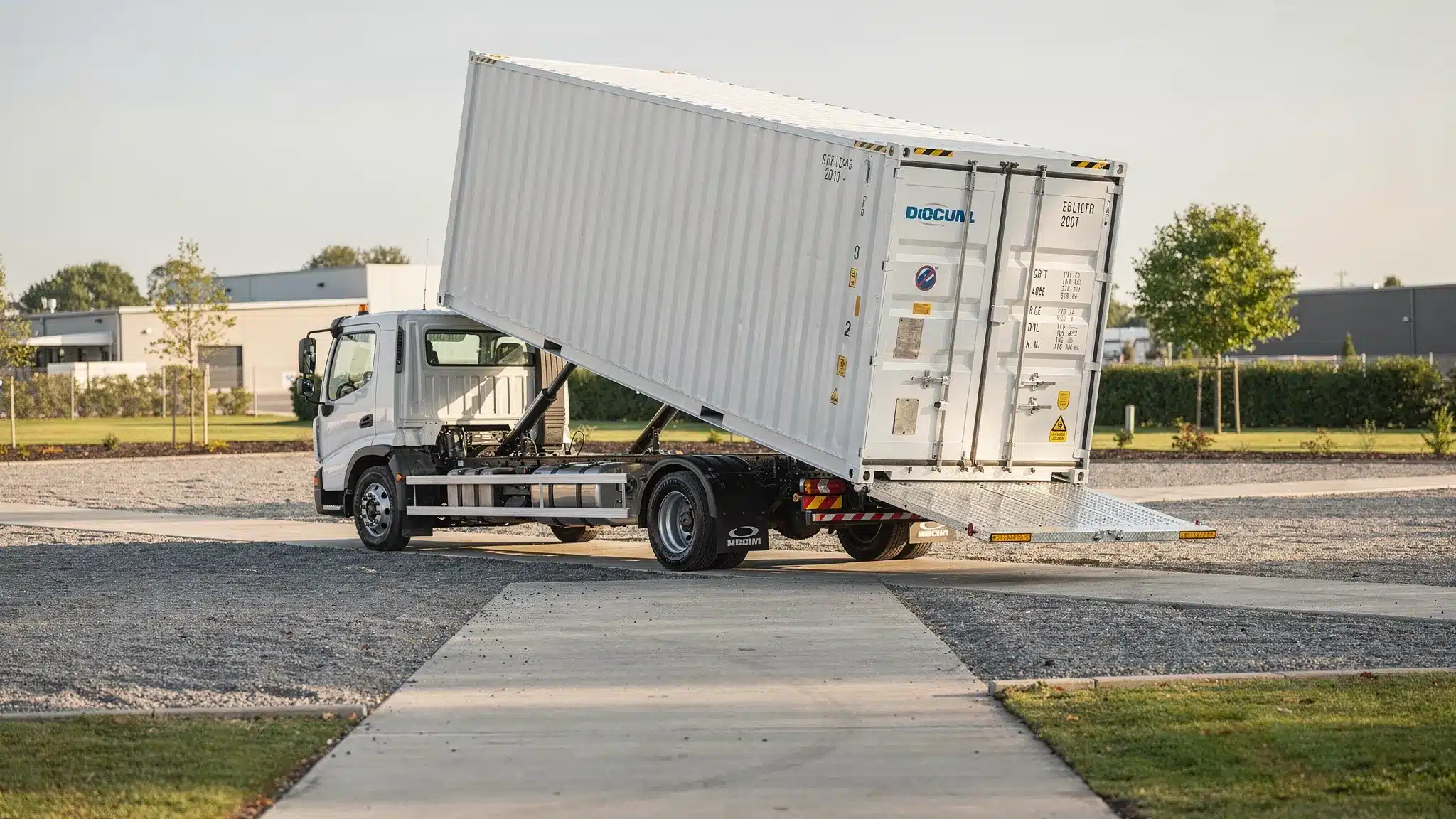 A delivery truck placing a 20ft cargo container on level gravel using a tilt-bed mechanism, with clear driveway access and ample turning space.