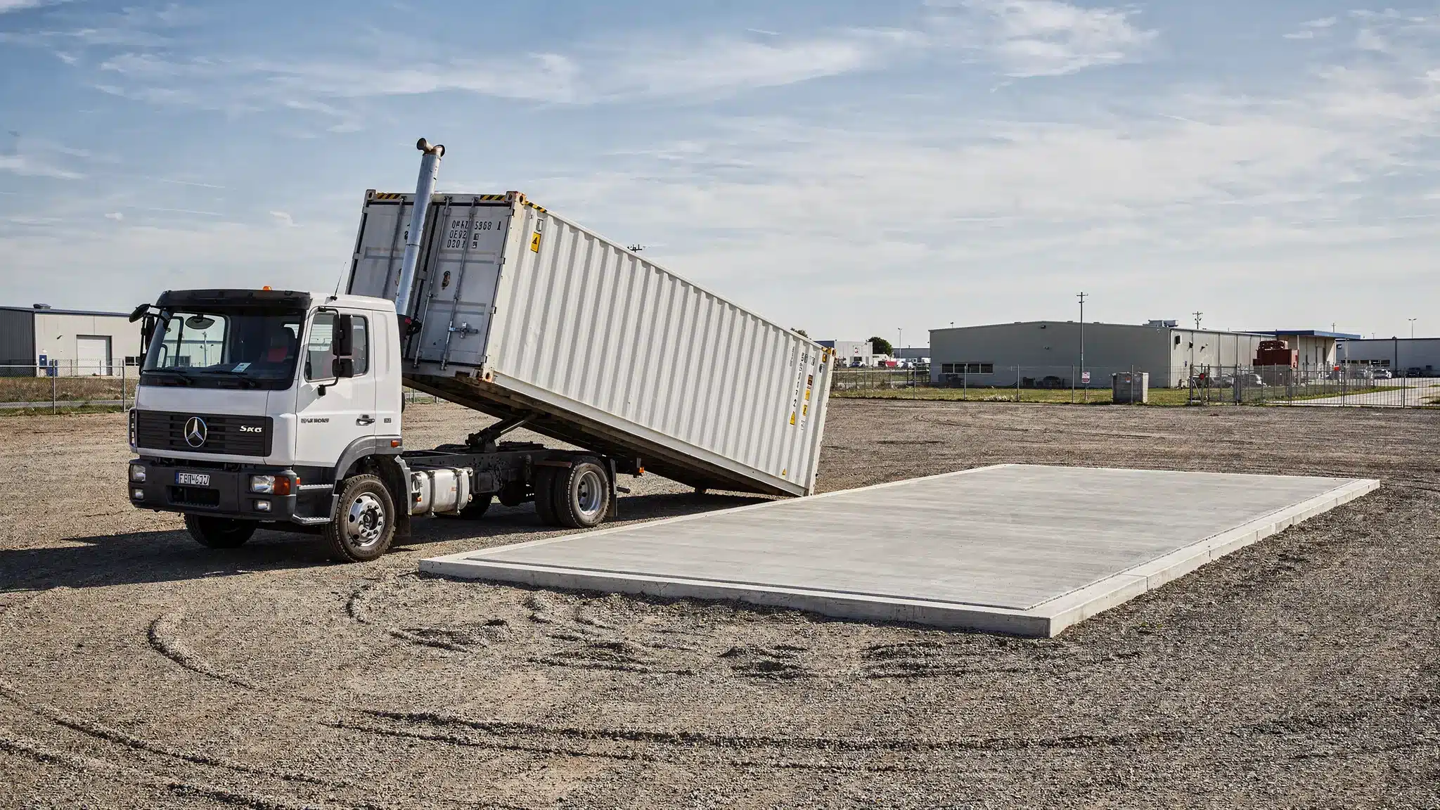 A delivery truck placing a shipping container at a prepared drop site, showing clear overhead space, level ground, and sufficient turning radius for placement.