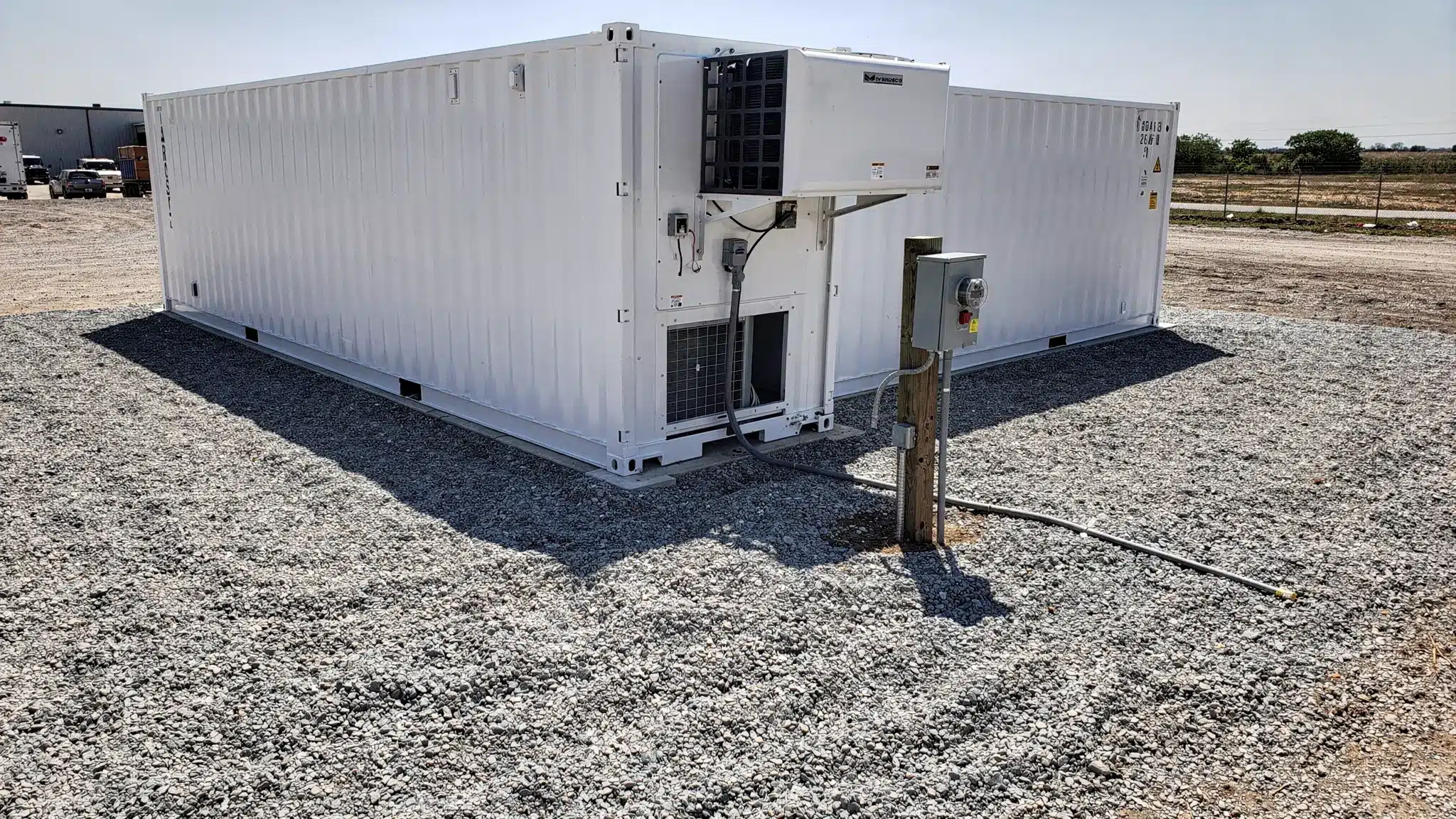 A refrigerated shipping container installed on a level gravel pad with clear space around the refrigeration unit end, showing proper airflow clearance and a nearby electrical disconnect on a post.