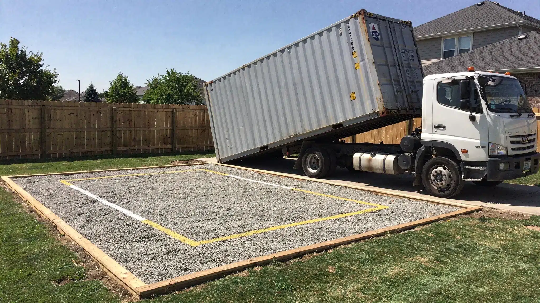 A shipping container being delivered on a tilt-bed truck to a prepared gravel pad on a residential property, with visible overhead clearance and a marked placement area near a fence line.