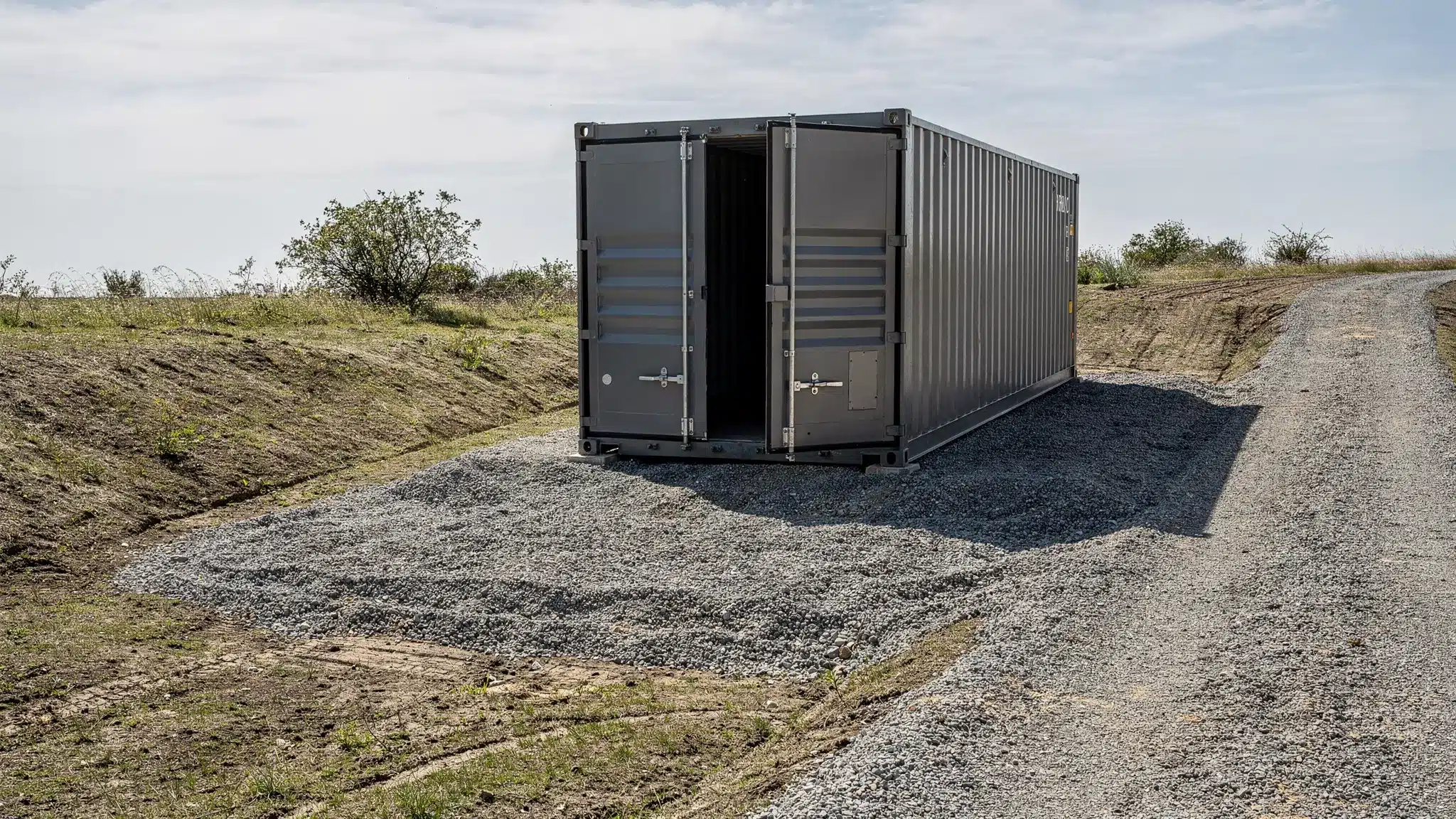 A shipping container placed on a compacted gravel pad with corner blocks, surrounded by a graded surface that slopes away for drainage, with clear door swing space and a visible approach path for a delivery truck.
