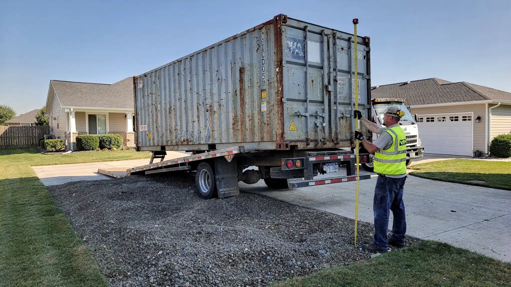 A used 10ft shipping container on a tilt-bed delivery truck being set down on a level gravel pad at a residential driveway, with clear overhead space and a tape measure showing site clearance checks.