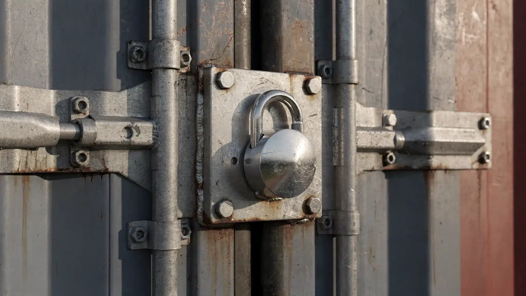 Secure Storage Containers for Sale: Locks, Doors, Upgrades 1 Close-up of a shipping container door with a welded steel lockbox installed over the hasp area and a heavy-duty puck lock engaged, showing reinforced door hardware and textured steel surface.