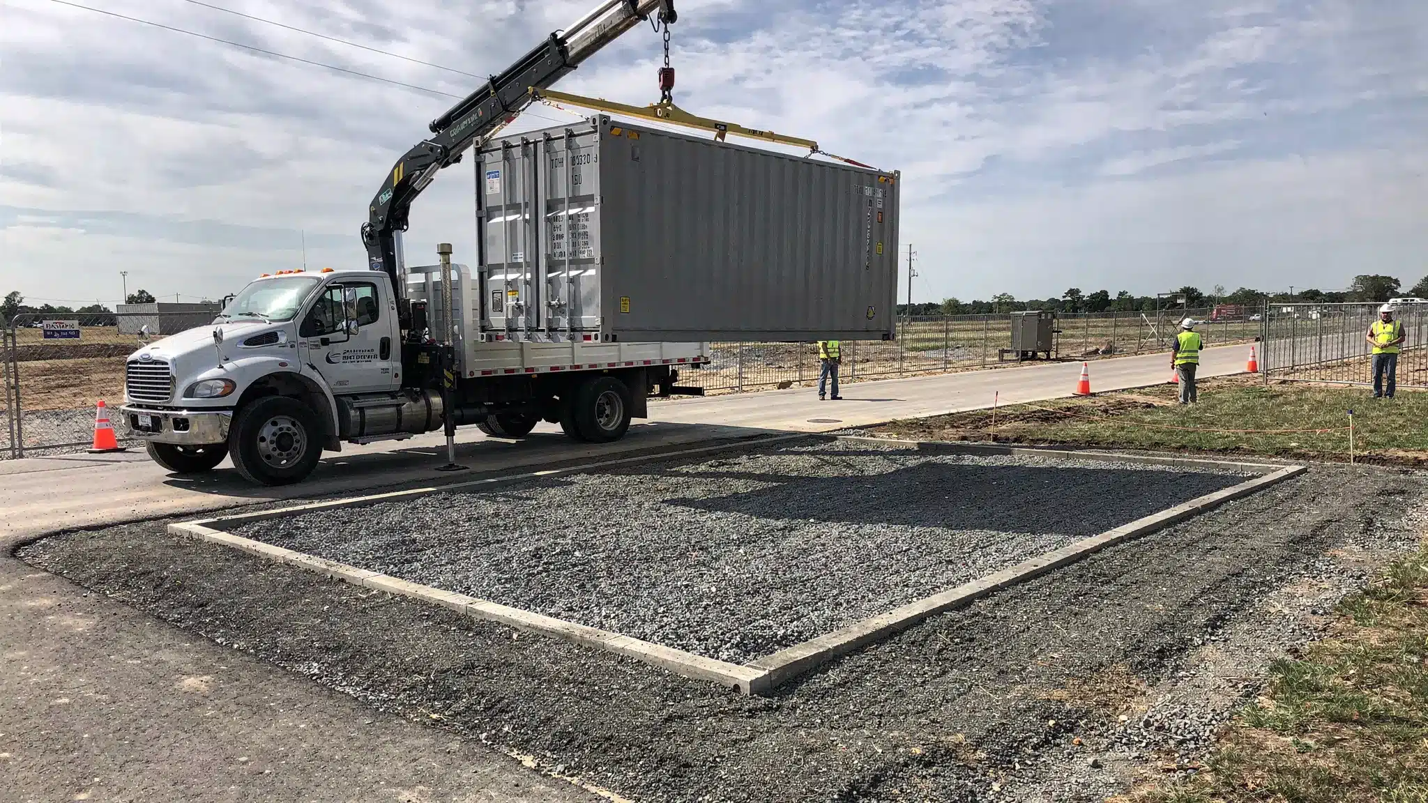 A delivery truck offloading a shipping container onto a prepared gravel pad at a U.S. jobsite with clear space, showing proper site access and placement.