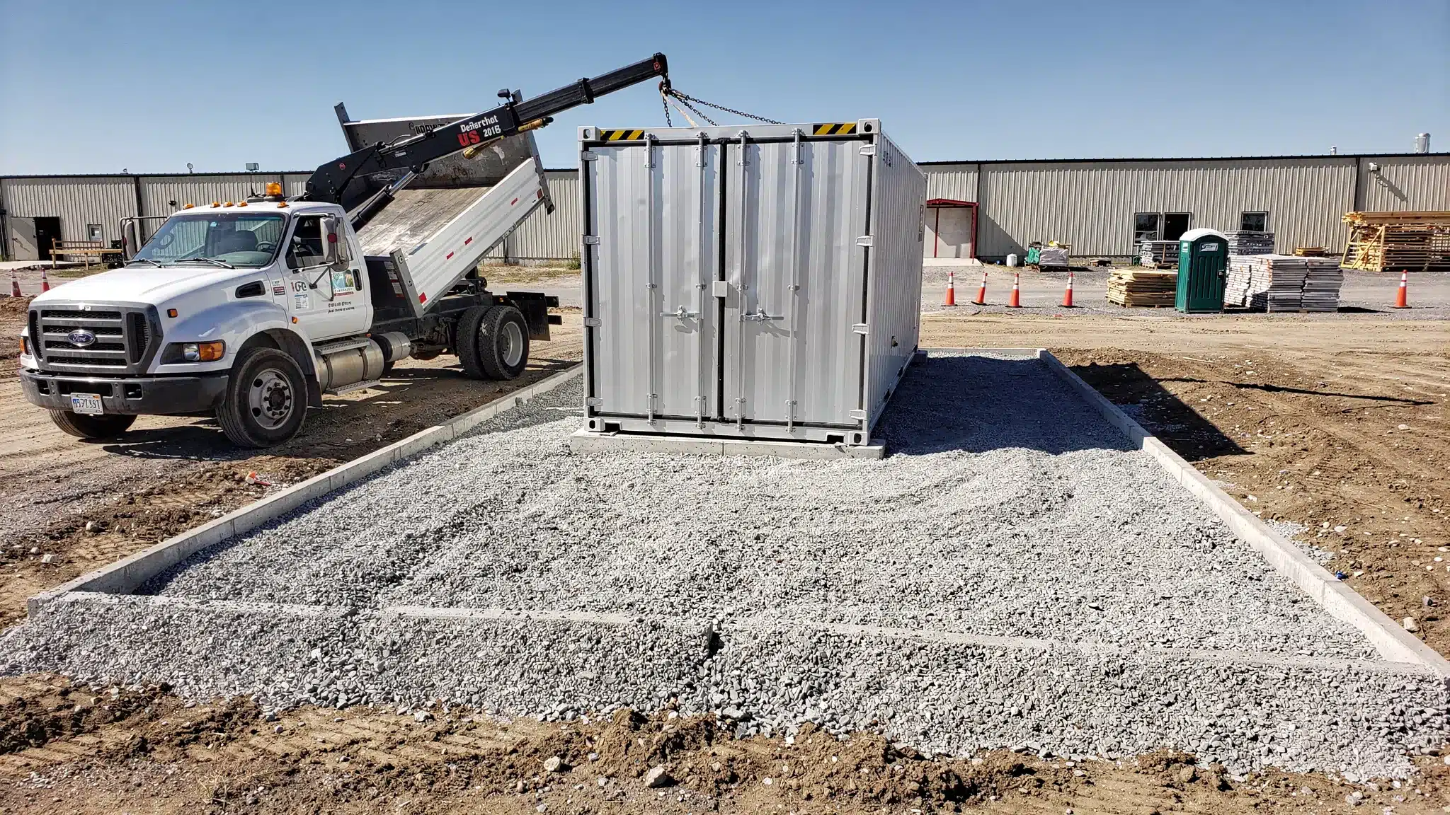 A delivery truck placing a 20ft shipping container on a prepared gravel pad at a U.S. job site, with clear space around the container for door access.