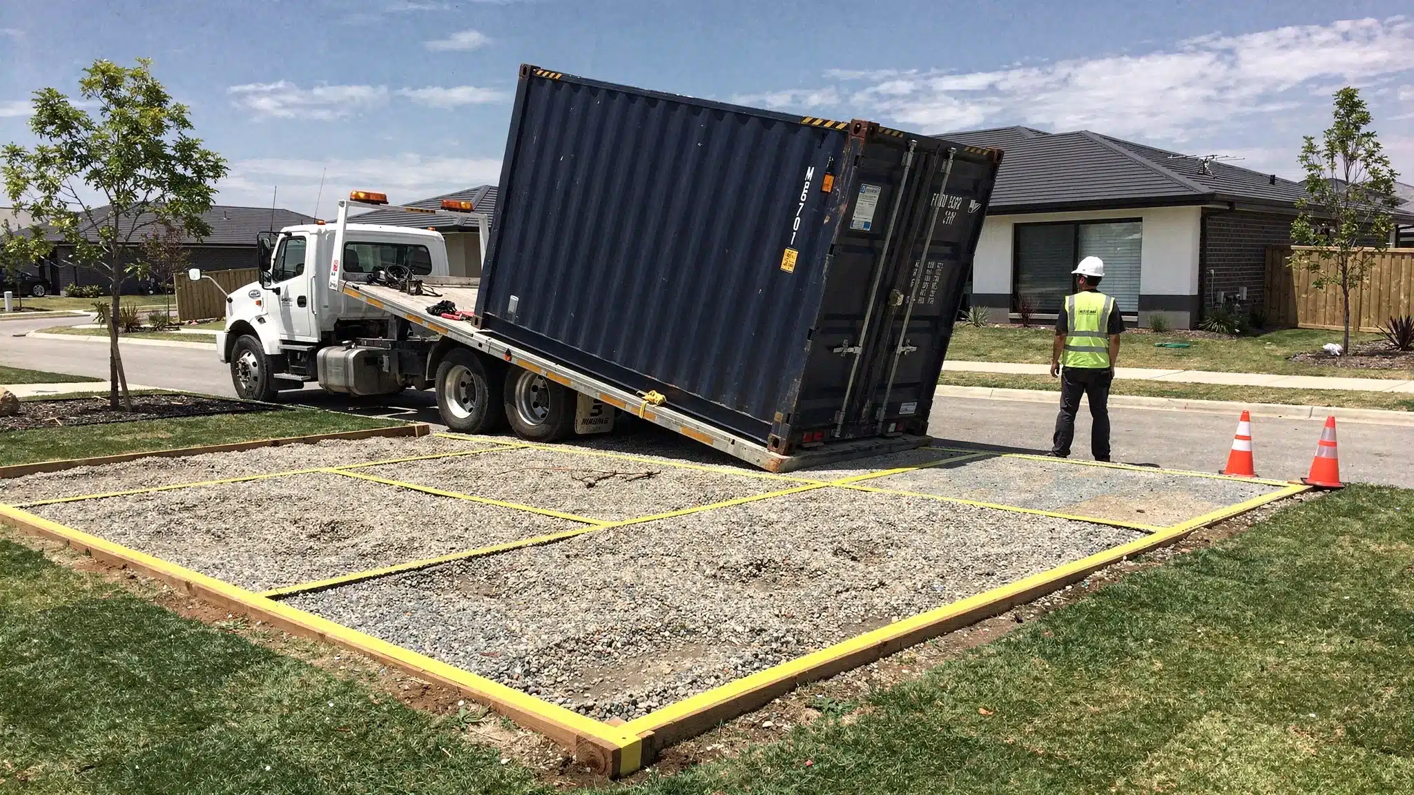 Rent to Own Sea Containers: Who It Fits and What to Watch 1 A shipping container being delivered on a tilt-bed truck onto a compact gravel pad at a residential property, with clear overhead space and a taped-off placement area.