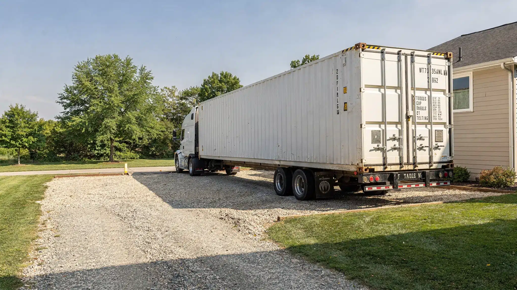 Purchase Shipping Containers Online: A Step-by-Step Guide 1 A shipping container delivery truck positioning a 20ft container onto a level gravel pad at a residential property, showing clear driveway access, overhead clearance, and the container doors facing outward.