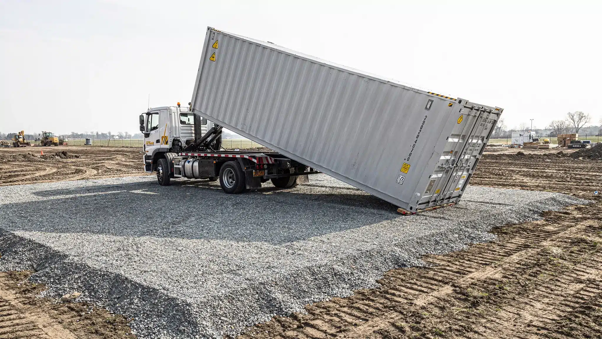 A tilt-bed delivery truck placing a 20ft shipping container onto a prepared gravel pad at a job site, with clear space around the truck and no overhead obstructions.