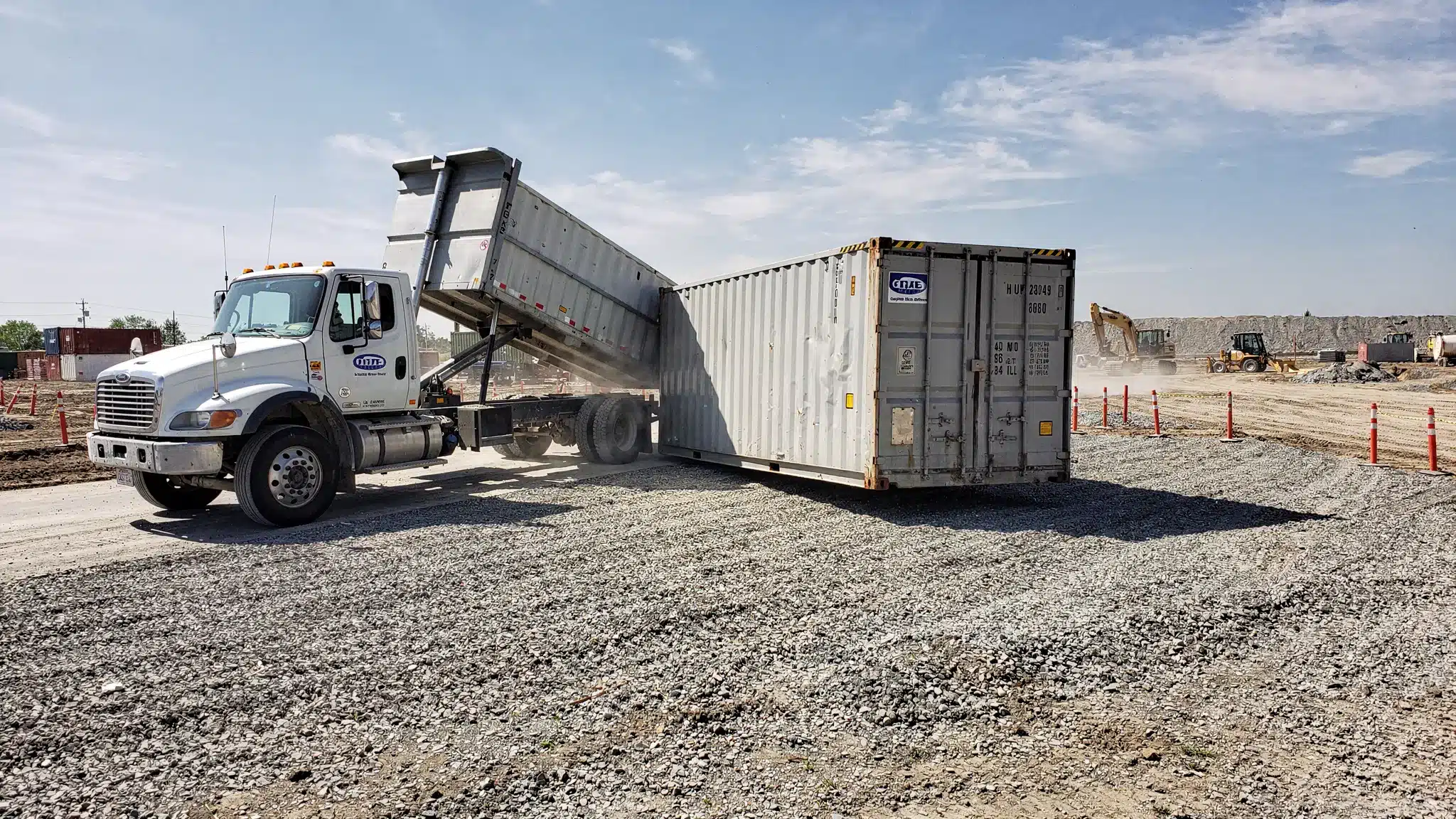 Seaworthy Containers for Sale: What “Cargo-Worthy” Means 2 A tilt-bed delivery truck unloading a 40ft shipping container onto a level gravel pad at a job site, with clear access space and the container doors facing outward.
