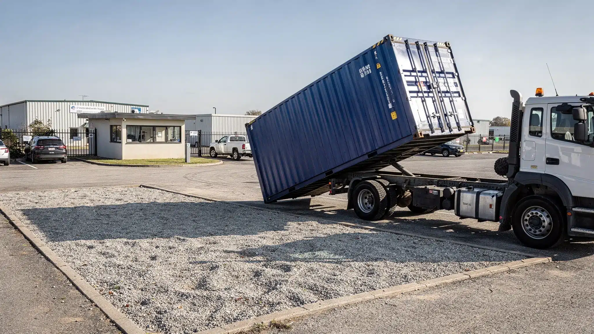 A tilt-bed truck delivering a blue shipping container to a prepared gravel pad at a small business yard, with clear space around the drop location and no overhead obstructions.