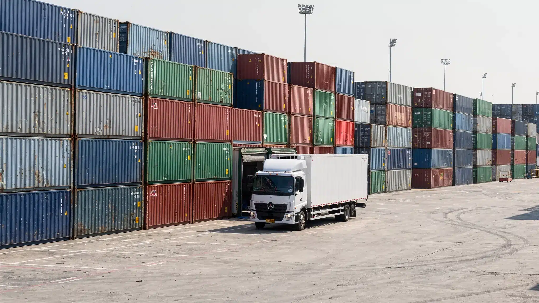 A shipping container yard with rows of 20ft and 40ft steel containers ready for delivery, with a delivery truck positioned near the front and open space for loading.